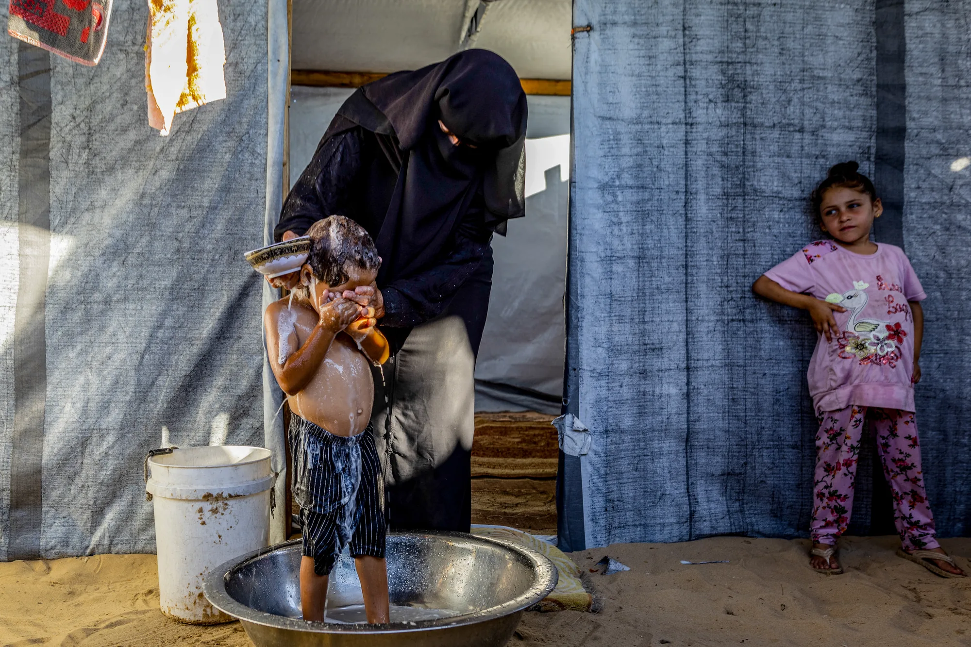 In a displacement camp in Gaza, a mother washes her son outside their shelter on a newly paved pathway. Restored access to water supply networks provides families with clean water for daily life and supports small-scale gardening, improving health, strengthening food security and helping restore everyday routines.